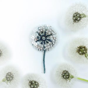 May include: A white and silver beaded dandelion brooch with a dark blue centre. The brooch is surrounded by white dandelions.