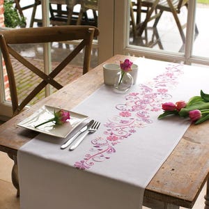 May include: White table runner with pink floral embroidery. The runner is on a wooden table with a white chair and a plate with a pink rose.