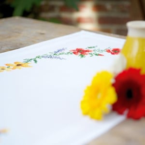 May include: A white embroidered table runner with a floral design featuring yellow, red, and purple flowers. The runner is on a wooden table with a yellow ceramic vase and red and yellow flowers.