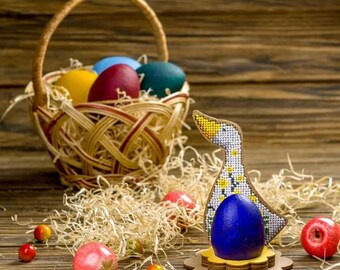Può includere: Un uovo di Pasqua blu si trova in un porta uova di legno a forma di anatra decorato con fiori gialli. L'anatra è in legno e presenta un motivo bianco e giallo. Il porta uova è su un tavolo di legno con un cesto di vimini pieno di uova di Pasqua colorate sullo sfondo.