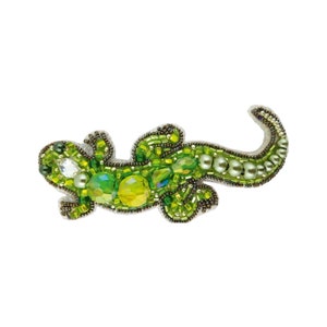 Puede incluir: Un broche de lagarto verde con cuentas verdes iridiscentes y blancas. El lagarto está en posición enroscada con la cola enroscada alrededor de su cuerpo.