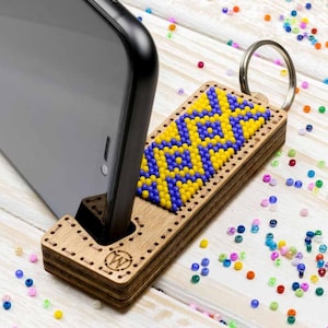 May include: A wooden phone stand with a beaded design in yellow and blue. The stand has a cutout for a phone and a key ring attached. The stand is sitting on a white surface with colorful beads scattered around it.