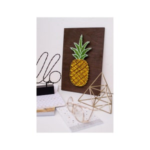 Op de afbeelding: Een houten wanddecoratie met een ananasontwerp van string art. De ananas is geel met groene bladeren en bevindt zich op een donkerbruine achtergrond.