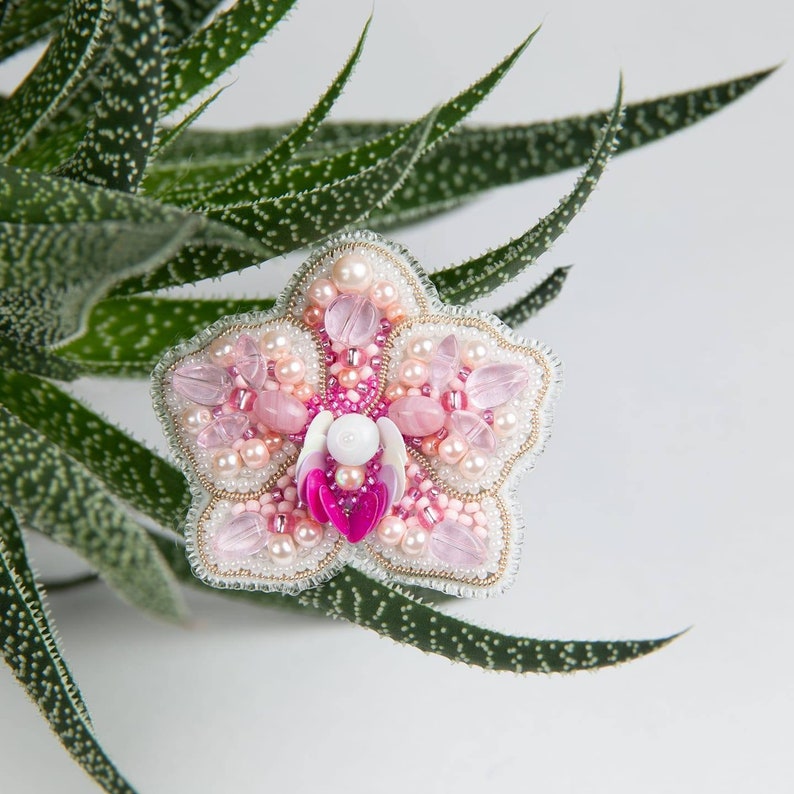 Pu&ograve; includere: Una spilla a forma di fiore in perle rosa e bianche con un centro madreperlaceo. La spilla &egrave; appoggiata su una pianta grassa verde.