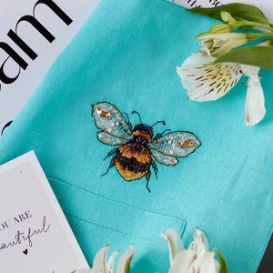 May include: A turquoise linen tote bag with a beaded bee embroidered on the front. The bag is decorated with white flowers.