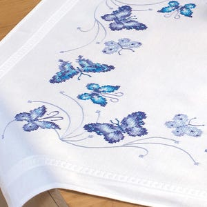 Könnte beinhalten: Weiße Tischdecke mit einem blauen Schmetterlings-Stickmuster. Die Schmetterlinge sind in einer Reihe entlang des Randes der Tischdecke angeordnet.