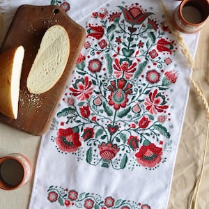 May include: White embroidered table runner with a floral design in red, green, and white. The runner is fringed and has a traditional folk art style. There are also some bread rolls, eggs, and a cup on the table.