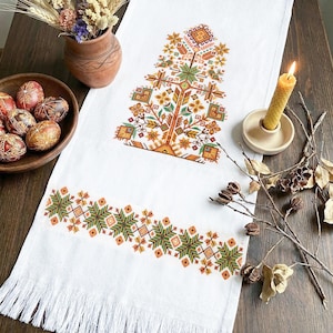 May include: White embroidered table runner with a tree design in shades of orange, green, and yellow. The runner has a fringed edge and is decorated with a geometric pattern. A wooden bowl with decorated eggs and a lit candle are also visible.