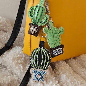 May include: Three cactus-shaped keyrings with green, black, and white beaded details. The keyrings are attached to a yellow handbag. The cactus keyrings have brown pots with white and blue accents.