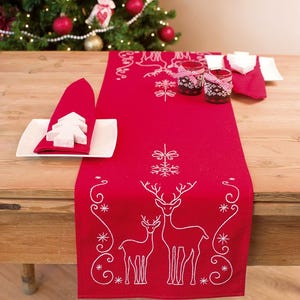 May include: Red table runner with white embroidered deer and snowflakes. The runner is on a wooden table with white napkins and glasses.