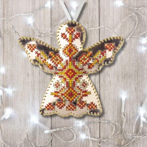 May include: A handcrafted angel ornament with a white body and embroidered wings. The wings are detailed with red, yellow, and brown cross-stitch patterns. A white ribbon is attached for hanging. The ornament is displayed against a wooden background with string lights.