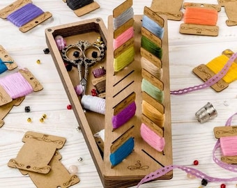 May include: A wooden box with a built-in thread organizer. The box has a hinged lid and a compartment for storing small items. The thread organizer has slots for holding spools of thread. The box is filled with colorful threads, beads, and other craft supplies.