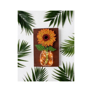May include: A string art sunflower with orange, yellow, and red petals is in a mason jar with white lights. The jar is on a brown wooden background.