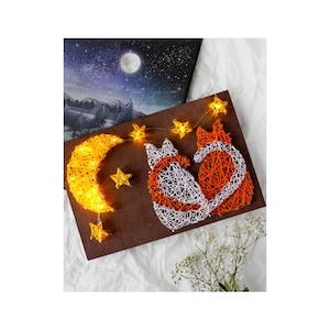 Puede incluir: Una pieza de arte de hilo que representa una luna creciente y dos zorros, iluminada con luces blancas cálidas. Los zorros son naranjas y blancos, sobre un fondo de madera oscura. Luces en forma de estrella están ensartadas en la parte superior, creando un objeto decorativo caprichoso.