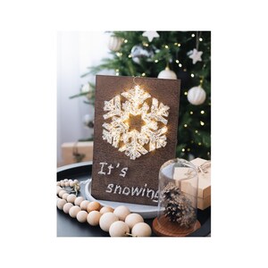 May include: A wooden sign with a string art snowflake design and the text "It's snowing". The snowflake is illuminated with white lights. The sign is sitting on a table with a wooden bead garland and a glass cloche with a pine cone inside.