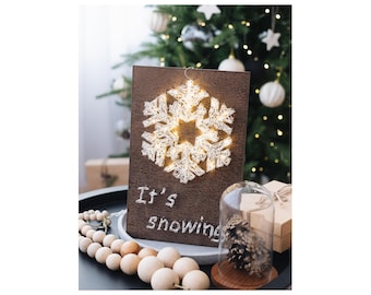 May include: A wooden sign with a string art snowflake design and the text "It's snowing". The snowflake is illuminated with white lights. The sign is sitting on a table with a wooden bead garland and a glass cloche with a pine cone inside.