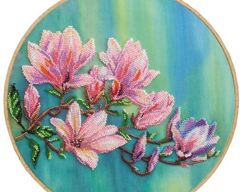 May include: A circular wooden embroidery hoop framing a piece of fabric with a hand-beaded design of pink and purple magnolia flowers on brown stems. The background fabric features a blended gradient of teal, blue, and light green colours.