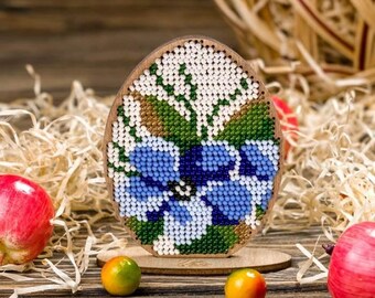 Può includere: Un ornamento per uovo di Pasqua in perline con un motivo floreale blu su un supporto in legno. L'uovo è decorato con perline blu, bianche e verdi.