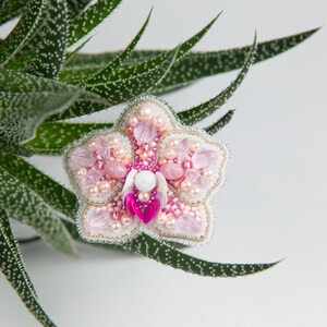 Pu&ograve; includere: Un broche a forma di orchidea, realizzato con perline rosa e bianche, con un centro di perla. Il broche &egrave; circondato da foglie verdi.