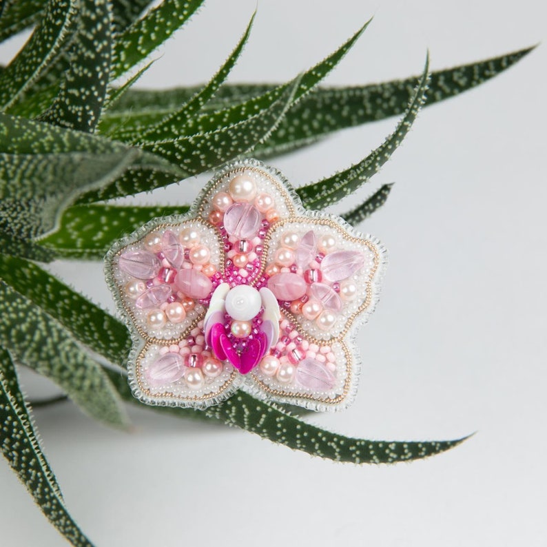 Pu&ograve; includere: Un broche a forma di orchidea, realizzato con perline rosa e bianche, con un centro di perla. Il broche &egrave; circondato da foglie verdi.