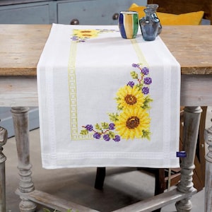 Op de afbeelding: Witte tafelrunner met geborduurde zonnebloemen en paarse bessen. De runner ligt op een houten tafel met een kruisvormig onderstel.