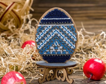 Può includere: Un ornamento a forma di uovo di Pasqua in perline blu e bianche su un supporto in legno. L'uovo è decorato con un motivo geometrico ed è circondato da paglia.
