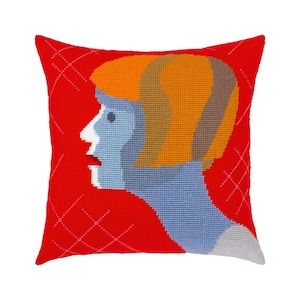 Puede incluir: Un cojín cuadrado con un diseño de punto de aguja que muestra el perfil de una cabeza. El fondo es rojo brillante con líneas de cuadrícula blancas bordadas. La figura tiene piel azul claro, cabello naranja y cuello blanco sobre el fondo rojo.