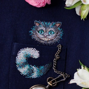 May include: A blue denim pocket with a white pocket watch and chain. The pocket has a cross-stitch design of the Cheshire Cat from Alice in Wonderland. The cat's face is stitched in blue and green thread, and the tail is stitched in a swirling pattern of blue, green, and white thread.