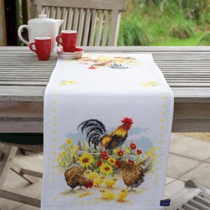 Puede incluir: Un camino de mesa blanco con un diseño de punto de cruz de un gallo y pollitos. El gallo es rojo, amarillo y negro. Los pollitos son amarillos. El diseño está rodeado de flores amarillas.