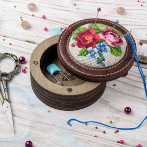 Puede incluir: Una caja redonda de madera con una tapa con bisagras. La tapa está decorada con un diseño de punto de cruz floral con flores rojas, rosas y azules. La caja está abierta y el interior está forrado de fieltro. Hay varias alfileres de costura clavados en el fieltro.