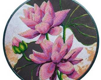 May include: A circular embroidery hoop containing a beadwork art piece featuring two large pink lotus flowers and a single green bud. The flowers are crafted with small beads in shades of pink, purple, and white, set against a dark background.