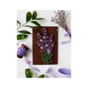 May include: A wooden plaque featuring a string art floral design in shades of purple and green. The artwork is mounted on a dark brown wooden board. A bundle of dried lavender, a spool of purple lace ribbon, and a watering can are also present.