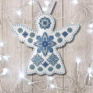 May include: A white, angel-shaped ornament with blue cross-stitch embroidery. The wings and body feature a detailed floral pattern. The ornament has a loop for hanging and is decorated with a string of white lights.
