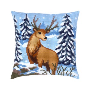 May include: A square throw pillow featuring a cross-stitch design of a brown deer standing in a snowy forest. The scene includes dark blue and white pine trees against a light blue sky with white snowflakes, creating a winter landscape.