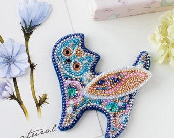 Op de afbeelding: Een blauwe, roze en gouden kralen broche met een grillig ontwerp. De kat zit met zijn staart om zijn lichaam gekruld. De broche is gemaakt met verschillende kralen, waaronder heldere, iriserende en ondoorzichtige kralen.