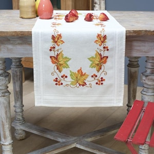 Könnte beinhalten: Ein weißer Tischläufer aus Leinen mit einem Kreuzstich-Design aus Herbstblättern in Grün-, Orange- und Rottönen. Der Läufer liegt auf einem Holztisch mit einem roten Klappstuhl im Vordergrund.