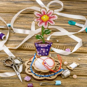 May include: A wooden flower pincushion featuring a pink flower with green leaves. The flower is in a purple pot with white polka dots. The pincushion has a white cushion and is surrounded by sewing supplies, including scissors, thread, and a thimble.