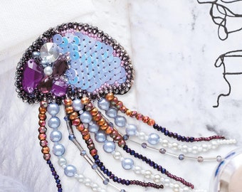 Op de afbeelding: Een broche in de vorm van een kwal, gemaakt van kralen, met een kleurenschema van paars, blauw en wit. De broche heeft een rond lichaam met een kralenrand.
