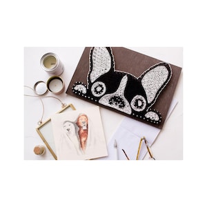 May include: A black and white string art piece of a French bulldog's face on a brown wooden background. The bulldog is looking at the camera with its tongue sticking out.