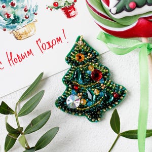 May include: A green beaded Christmas tree ornament with red, gold, and blue embellishments. The ornament is on a white surface with green leaves.
