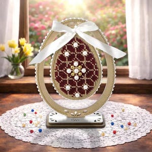 Puede incluir: Un huevo de Pascua decorativo con un diseño de cuentas, en un marco de madera. El huevo presenta un fondo burdeos con cuentas blancas y doradas. Una cinta blanca está atada en la parte superior. El huevo se exhibe sobre un soporte de madera.