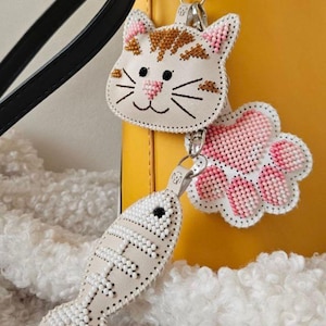 May include: Three beaded cat-themed keychains. One is a cat face with brown and white details, another is a pink paw print, and the third is a fish. All have silver-tone clasps and are attached to a yellow bag.