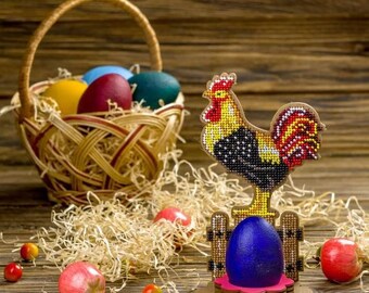 Può includere: Una statuetta di gallo colorata con perline in piedi su una base di legno con un uovo di Pasqua blu. Il gallo tiene una recinzione con un design rosso, giallo e nero. Un cesto di vimini pieno di uova di Pasqua colorate si trova sullo sfondo.
