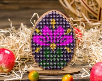 Può includere: Decorazione a forma di uovo di Pasqua in legno con un motivo floreale di perline nei toni del viola, rosa, giallo e verde. L'uovo è posto su un supporto di legno, circondato da paglia decorativa, uova rosse e piccoli oggetti rotondi colorati.
