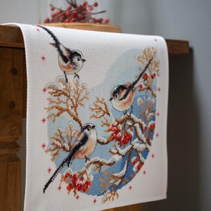 May include: White table runner with a cross-stitch design of three birds on a snowy branch with red berries. The birds are brown, white, and black.