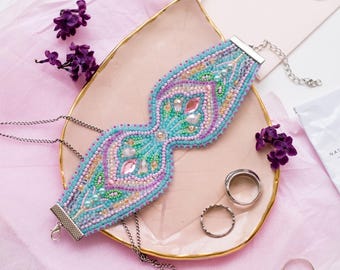 May include: A wide, intricately beaded choker necklace featuring a symmetrical pattern of light blue, lavender, and cream seed beads. The design includes central pink and clear faceted crystals, finished with silver-toned metal end caps and a chain.