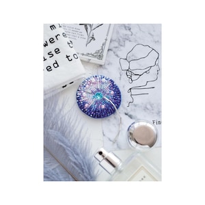 May include: A blue and pink beaded round ornament with a silver hook. The ornament has a blue and turquoise centre with white and pink beads surrounding it. The ornament is on a white marble surface with a feather and a bottle of perfume.