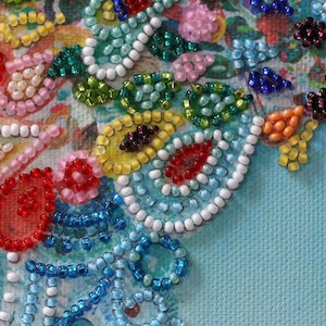 May include: Close-up of a beaded design with various colors, including red, white, blue, green, and pink. The beads are arranged in a pattern of circles, ovals, and swirls. The background is a light blue fabric.