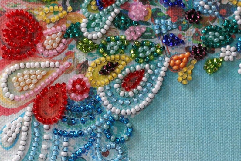 May include: Close-up of a beaded design with various colors, including red, white, blue, green, and pink. The beads are arranged in a pattern of circles, ovals, and swirls. The background is a light blue fabric.