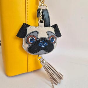 May include: A pug-shaped bag charm featuring a beige face with black ears and a black muzzle. The eyes are detailed with blue and brown stitching. A gold-tone metal chain connects the charm to a black strap, and a beige tassel hangs from the bottom.